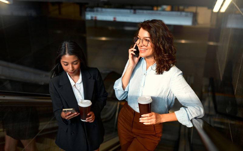 A team of successful business women of different races discuss strategy, drink coffee and use the phone on their way to the office. Diverse women going up the escalator.